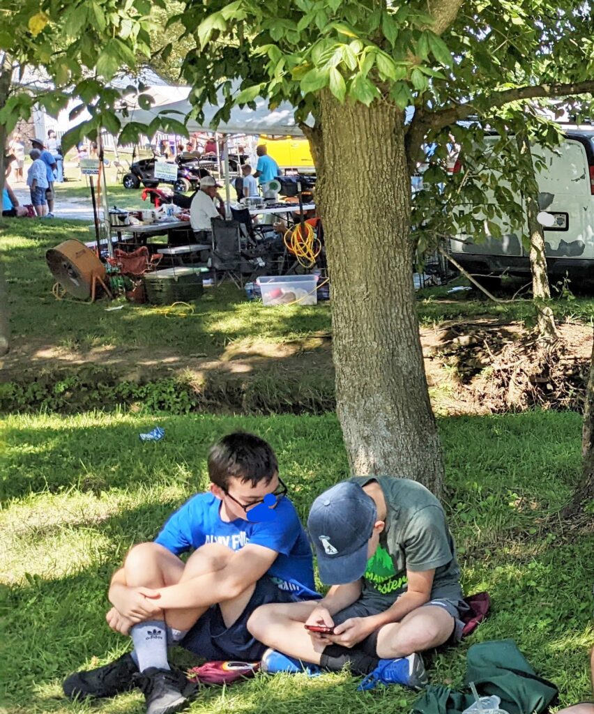 two boys sit under a tree looking at a phone with tents in background